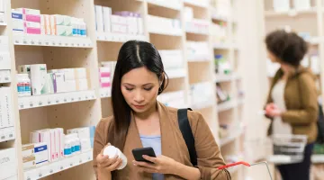 A young woman at a pharmacy inspects a bottle of supplements in her hand while also looking up supplement information on her cell phone.